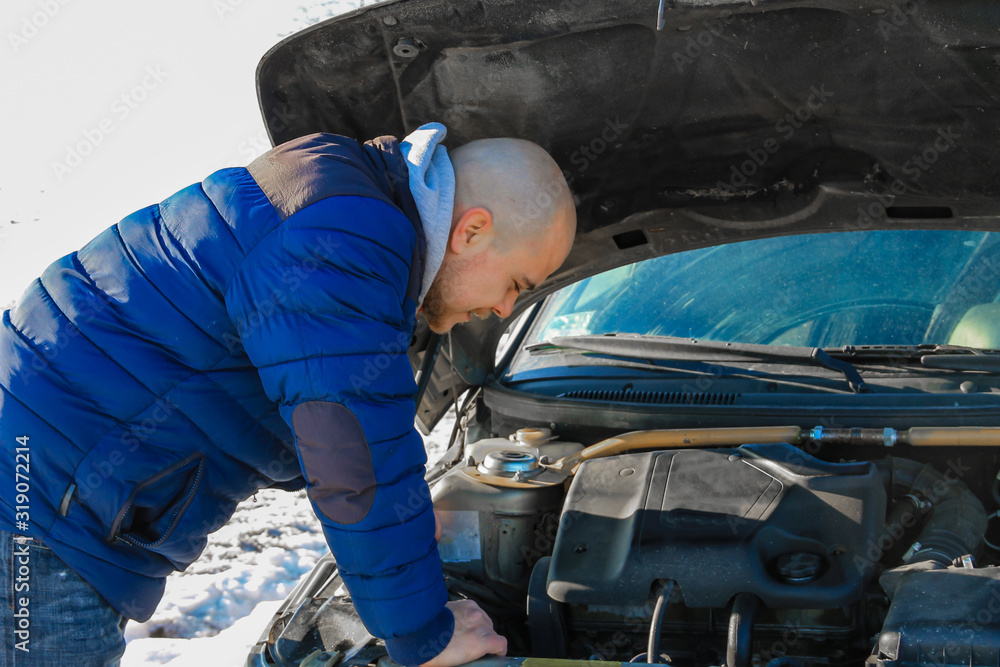 Young man looking under the hood of broken car in cold winter day Young man looking under the hood of broken car in cold winter day