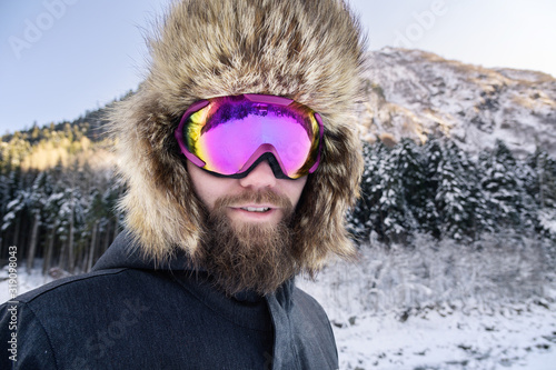 Close-up portrait of a bearded happy snowboarder skier in a ski mask with goggles and a fur big old-school hat on a background of a winter snowy mountains