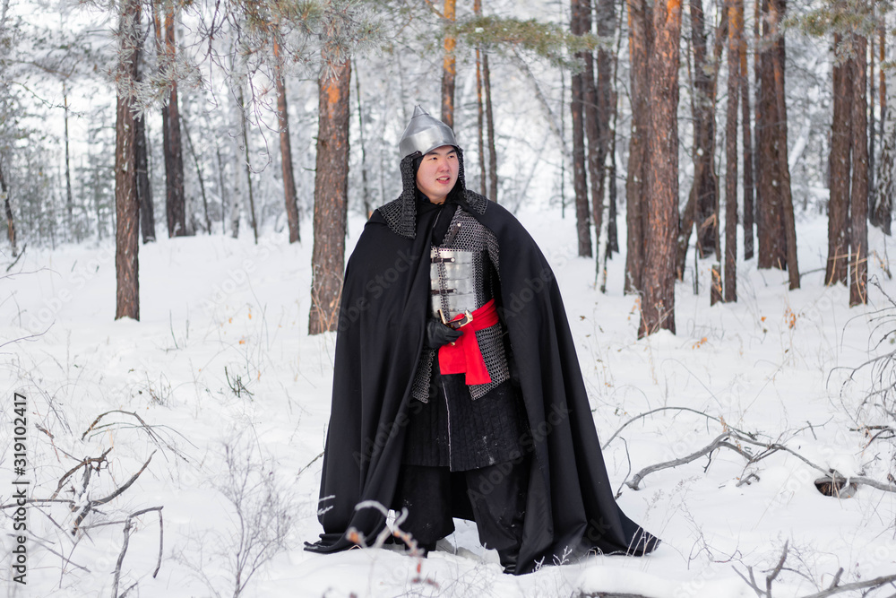 Medieval warrior in armor, helmet, black cloak with a saber in his hands in the winter in the forest. Medieval warrior in armor, helmet, black cloak with a saber in his hands in the winter in the forest.