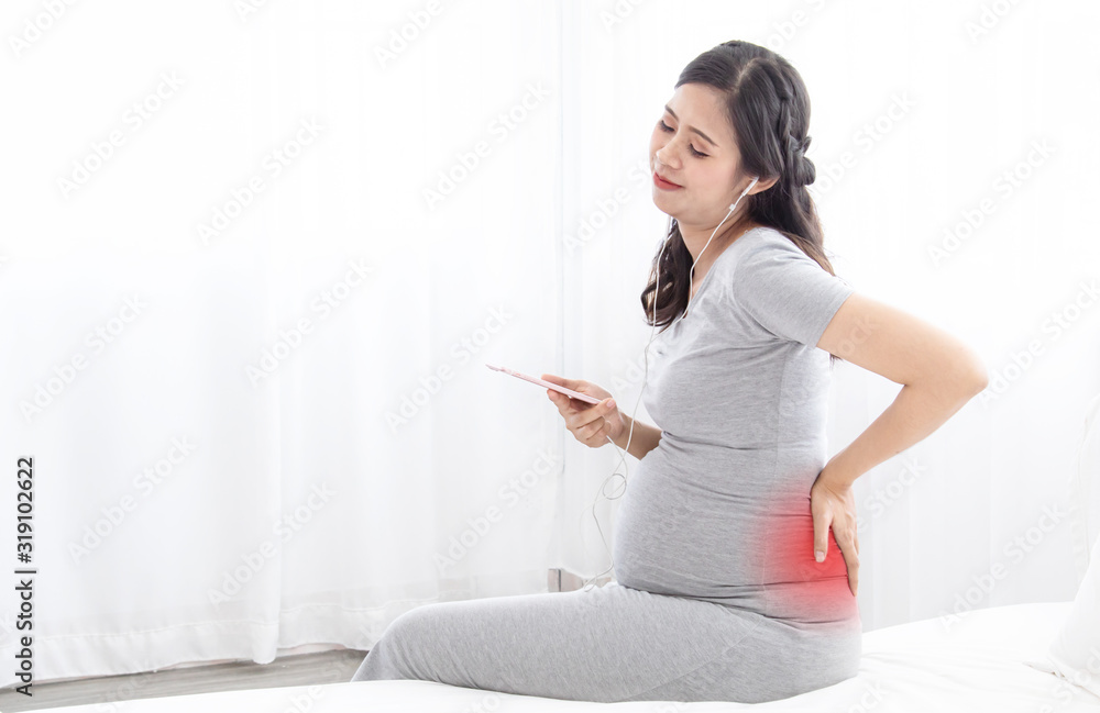 Beautiful pregnant Asian woman get sick strong pain backache sitting on bed while listen to music to relieve painful or call doctor advisory, nearly birth pregnant woman with red pain point concept Beautiful pregnant Asian woman get sick strong pain backache sitting on bed while listen to music to relieve painful or call doctor advisory, nearly birth pregnant woman with red pain point concept