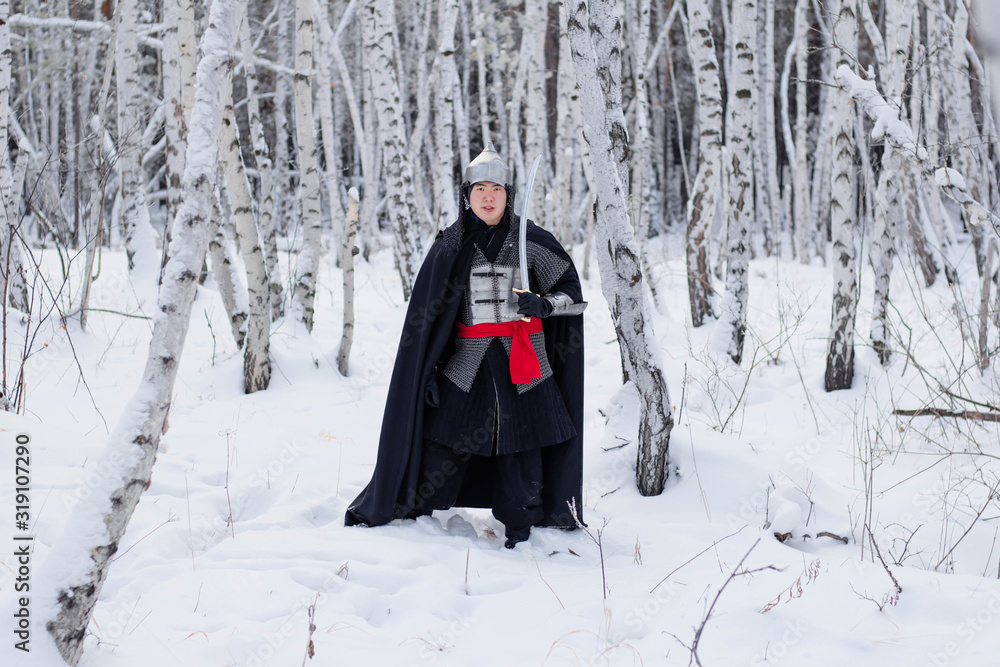 Medieval warrior in chain mail, helmet, black cloak with a saber in his hands against the background of a winter birch forest. Medieval warrior in chain mail, helmet, black cloak with a saber in his hands against the background of a winter birch forest.