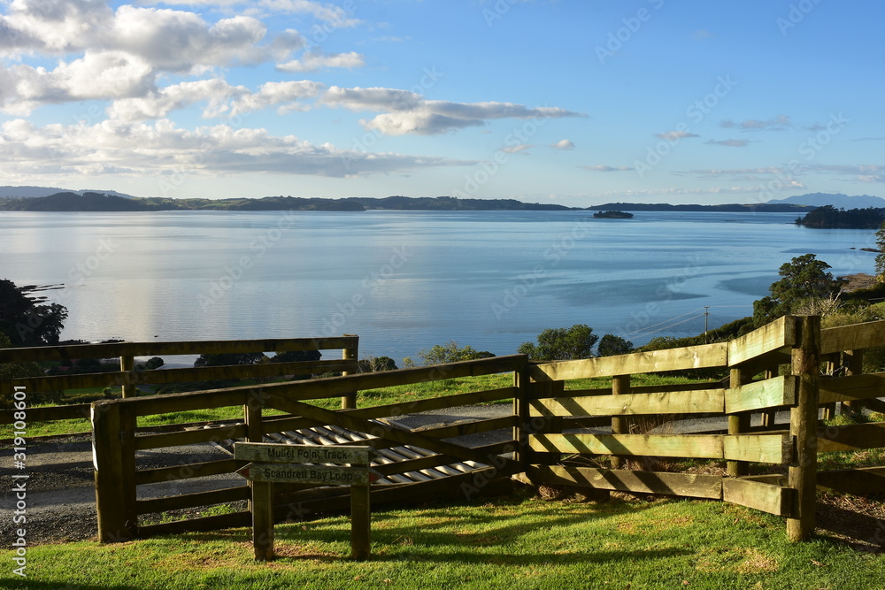 Wooden paddock with signs marking hiking trails along coast of calm bay near Auckland.