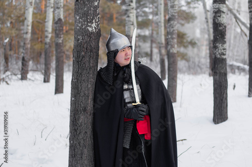 A medieval warrior in chain mail armor, a helmet and a black cloak with a saber in his hands, stands aside to a tree. Against the background of winter forest and snow.