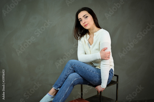 Portrait of a pretty young brunette woman in a white sweater and blue jeans sits on a stand. The concept of a cute girl on a gray background. Smiling, showing hands with emotions.