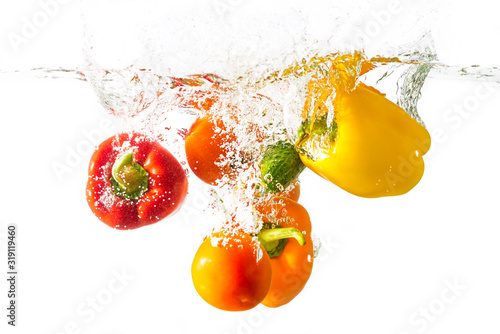 Three bell peppers, tomato and cucumber fall into the water with splashes, isolated on a white background.