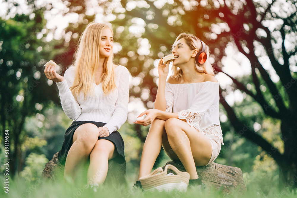 Young attractive caucasian buddy woman sitting in a park, eat sandwich togetherness and snack in brown handmade basket on lawn. Outdoor activity in summer to relax outside with friend,relationship Young attractive caucasian buddy woman sitting in a park, eat sandwich togetherness and snack in brown handmade basket on lawn. Outdoor activity in summer to relax outside with friend,relationship