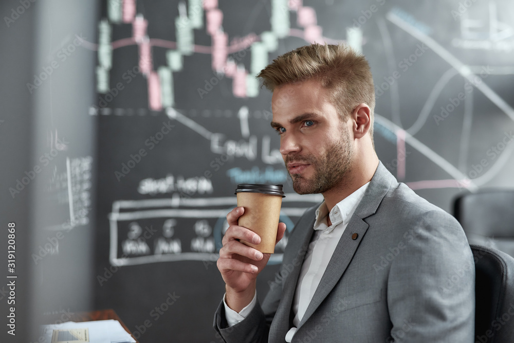 Experience the power of the broking. Close up portrait of successful young trader looking aside and drinking coffee while sitting in the office. Blackboard full of charts in background. Experience the power of the broking. Close up portrait of successful young trader looking aside and drinking coffee while sitting in the office. Blackboard full of charts in background.