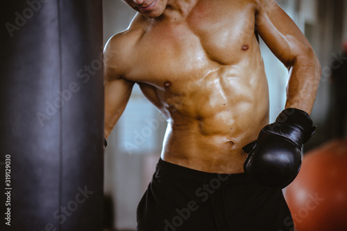 A sporty man trains a boxing training at gym,practice is showing strong six-pack muscles on front body while punching his hand to sandbag. Boxer bodybuilding exercise in studio with trainer instructor