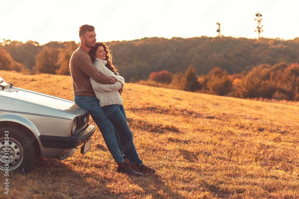 Beautiful young couple enjoying time together outdoor sitting on meadow leaning on old fashioned car embracing each other.