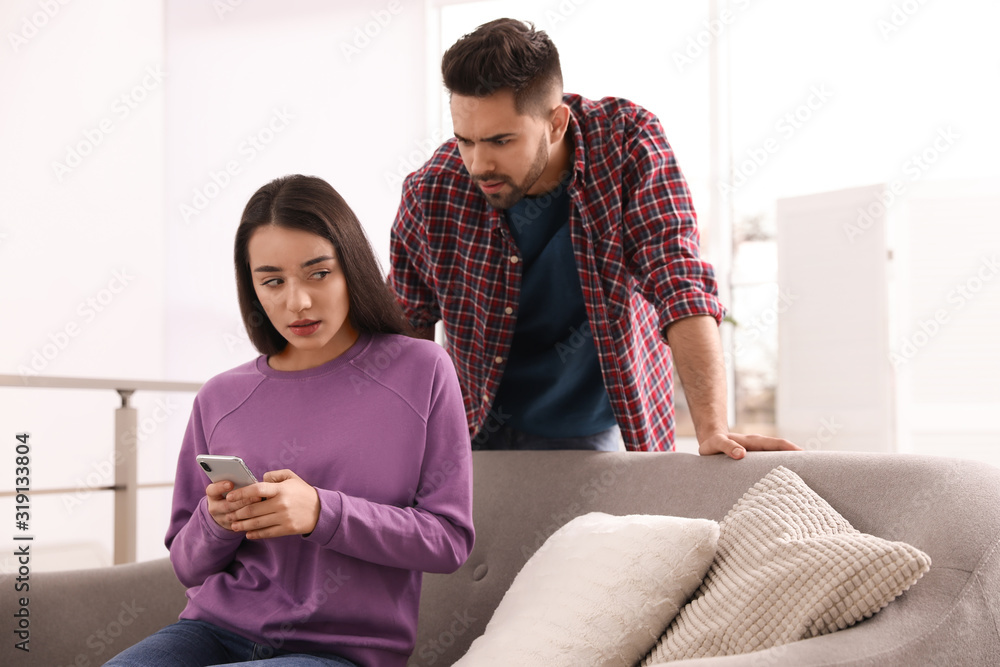 Distrustful young man peering into girlfriend's smartphone at home Distrustful young man peering into girlfriend's smartphone at home