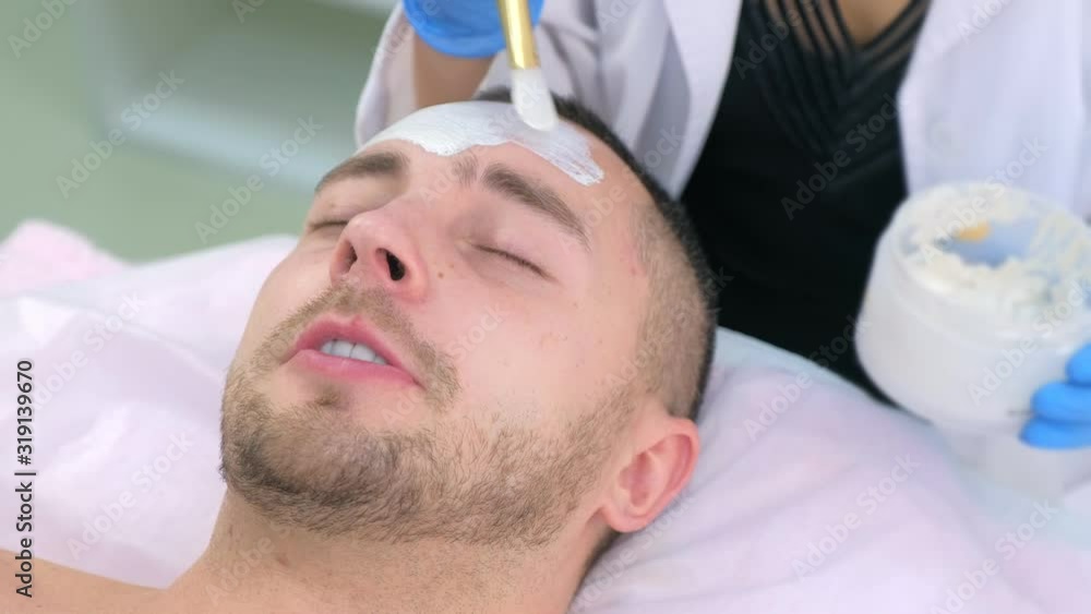 Cosmetologist is applying white mask on man client face using brush in beauty clinic. Portrait of guy closeup. Beautician making beauty facial skincare procedure to patient. Beauty industry concept.