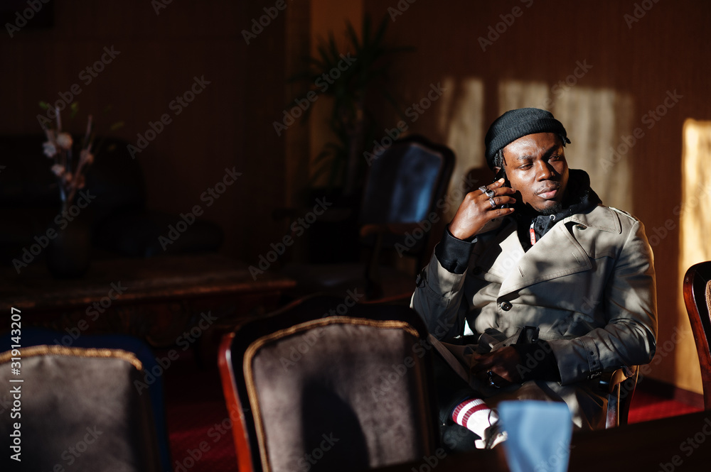 Handsome african american man posing inside room with sunlight shadows in black hat and beige coat, speaking on mobile phone. Handsome african american man posing inside room with sunlight shadows in black hat and beige coat, speaking on mobile phone.