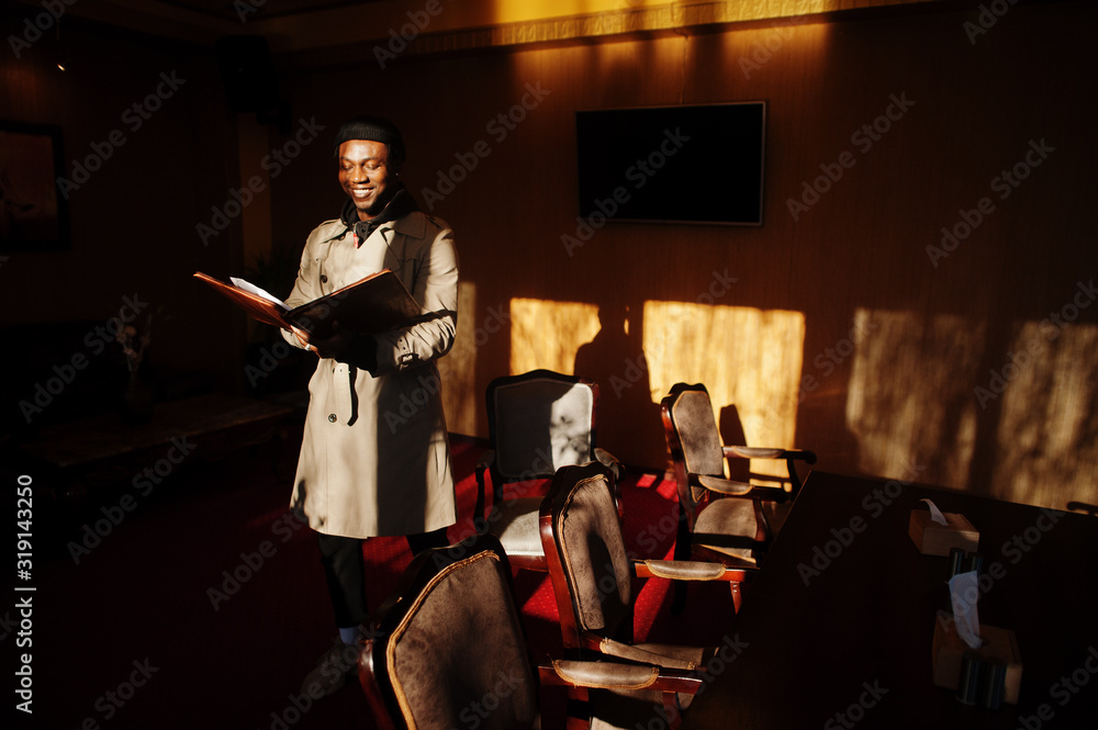 Handsome african american man posing  inside room with sunlight shadows in black hat and beige coat.