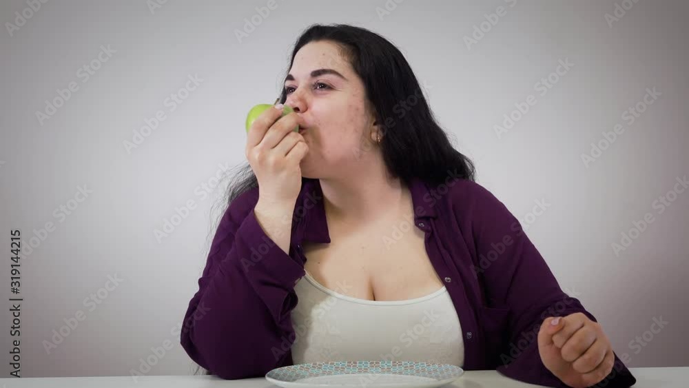 Portrait of cheerful obese Caucasian woman sitting at the table and eating apple. Smiling brunette girl eats healthy low-calorie food. Healthy lifestyle, dieting, healthcare. Portrait of cheerful obese Caucasian woman sitting at the table and eating apple. Smiling brunette girl eats healthy low-calorie food. Healthy lifestyle, dieting, healthcare.