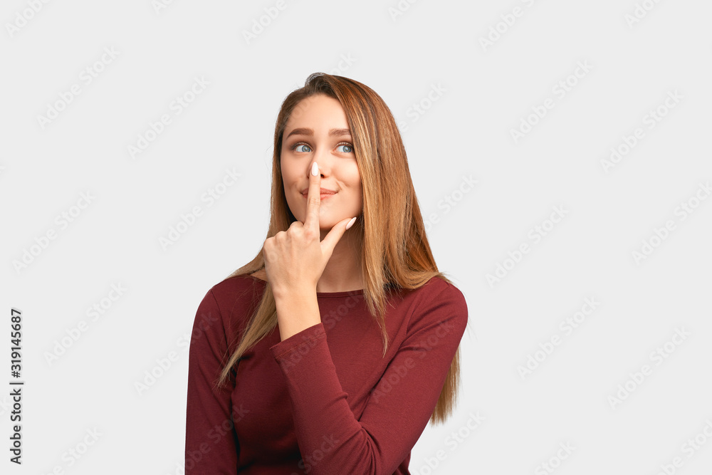 Funny girl holding index finger on nose, smiling sweetly. Beautiful young woman with long hair, clean smooth skin isolated on white background in Studio with an empty space for text and advertising. Funny girl holding index finger on nose, smiling sweetly. Beautiful young woman with long hair, clean smooth skin isolated on white background in Studio with an empty space for text and advertising.