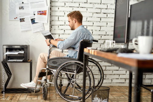 Portrait of young male office worker in a wheelchair reading some document while sitting near printer at his workplace in the bright modern office