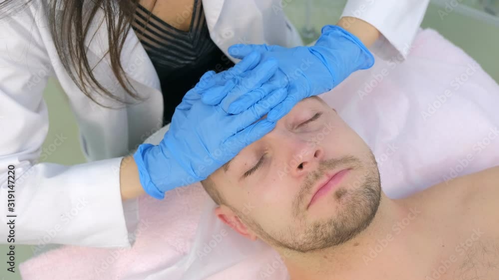 Cosmetologist in gloves is applying cream on client's man face massaging skin, top view. Beautician making beauty facial skincare procedure in clinic, portrait of guy patient. Beauty industry concept. Cosmetologist in gloves is applying cream on client's man face massaging skin, top view. Beautician making beauty facial skincare procedure in clinic, portrait of guy patient. Beauty industry concept.