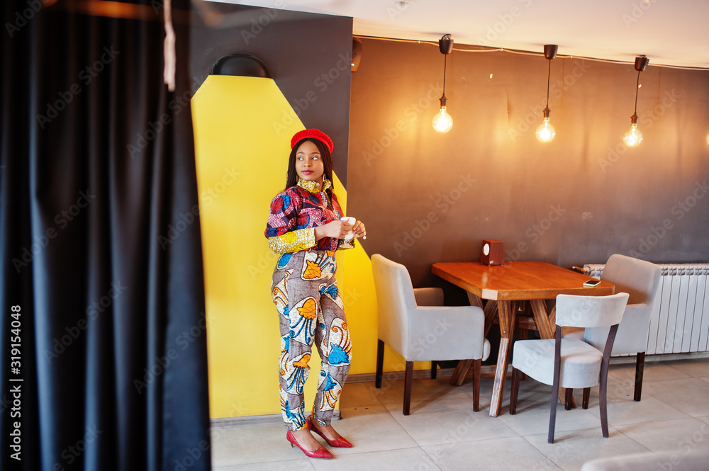 Enthusiastic african american woman in trendy coloured outfit with red beret chilling in cozy cafe with cup of hot latte in hands.