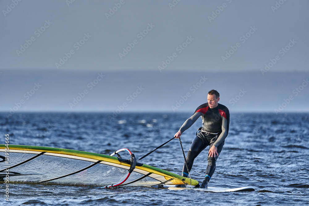 Naklejka premium A male athlete is interested in windsurfing. He moves on a Sailboard on a large lake on an autumn day.