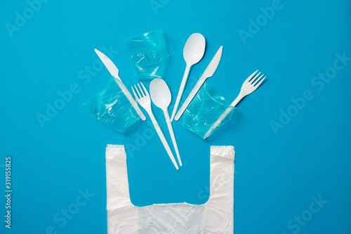 White single-use plastic knives, spoons, forks and bag on a blue background. Say no to single use plastic. Environmental, pollution concept.