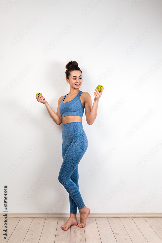 The concept of healthy eating. A young Caucasian woman in lilac sportswear, with a toned figure, holding green apples. White background. Copy space. Vertical The concept of healthy eating. A young Caucasian woman in lilac sportswear, with a toned figure, holding green apples. White background. Copy space. Vertical
