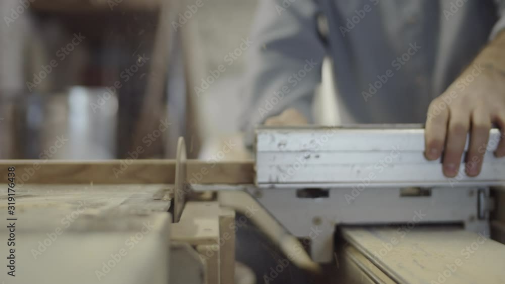 Close-up shot of hands of unrecognizable carpenter firmly holding piece of wood against blade while cutting it on table saw in workshop, and sawdust flying around Close-up shot of hands of unrecognizable carpenter firmly holding piece of wood against blade while cutting it on table saw in workshop, and sawdust flying around