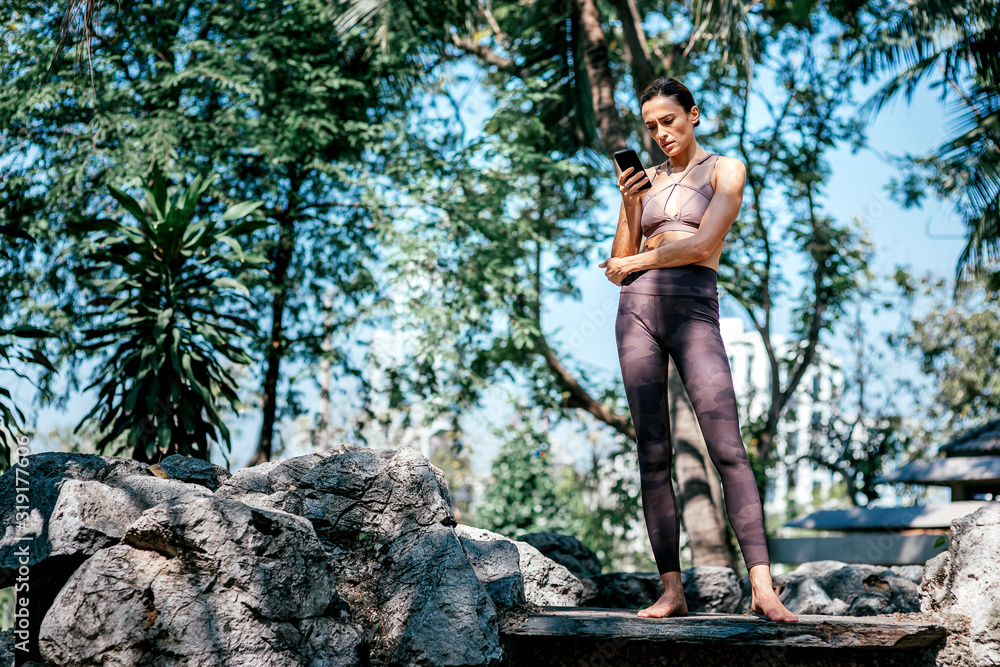 Add yoga in your life. Full-length shot of caucasian woman standing and looking at her smartphone while going to practice yoga outdoors, in the nature park. Healthy lifestyle and relaxation concept Add yoga in your life. Full-length shot of caucasian woman standing and looking at her smartphone while going to practice yoga outdoors, in the nature park. Healthy lifestyle and relaxation concept