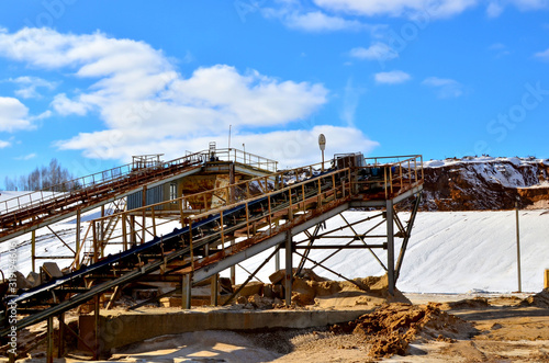 Mining plant in the open sand pit for the production of crushed stone, sand and gravel. View of the crushing machines and conveyor belts against the blue sky with clouds