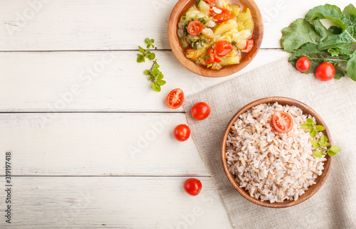 Unpolished rice porridge with stewed vegetables and oregano in wooden bowl on a white wooden background. Top view, copy space.
