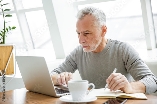 Photo of focused mature man making notes and working with laptop