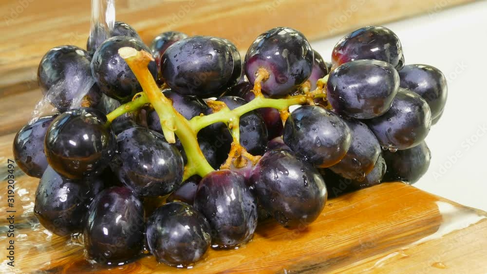 Close POV shot of clean water pouring over a bunch of freshly picked black grapes, on a pine draining board next to a sink. Close POV shot of clean water pouring over a bunch of freshly picked black grapes, on a pine draining board next to a sink.