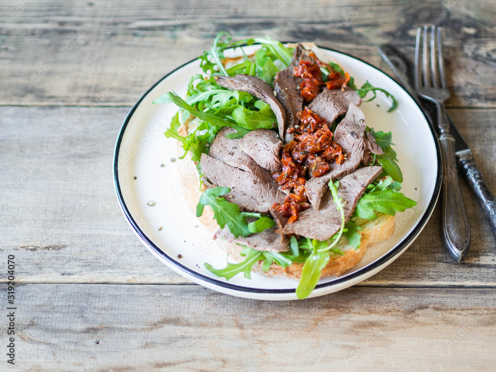 Large toast on homemade bread with soft cheese, arugula and slices of roast beef with sun-dried tomatoes on top on white plate on wood rustic background. Copy space. Large toast on homemade bread with soft cheese, arugula and slices of roast beef with sun-dried tomatoes on top on white plate on wood rustic background. Copy space.