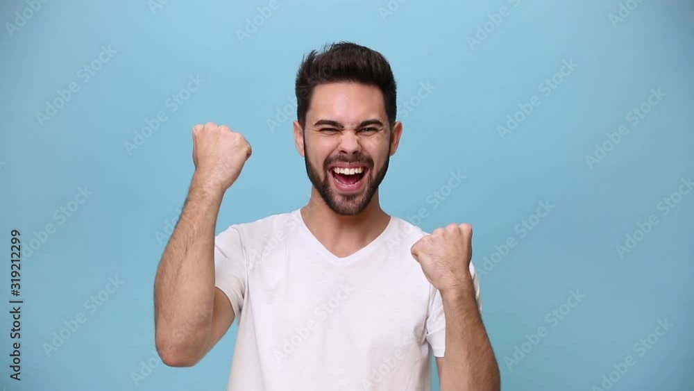 Handsome unshaven bearded young guy 20s wearing white t-shirt isolated over pastel blue background in studio. People sincere emotions lifestyle concept. Looking at camera doing winner gesture, say Yes Handsome unshaven bearded young guy 20s wearing white t-shirt isolated over pastel blue background in studio. People sincere emotions lifestyle concept. Looking at camera doing winner gesture, say Yes