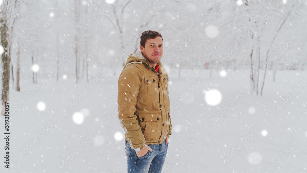 Portrait of young man in jacket and jeans in winter season. Handsome man stands and squint in winter park. Fluffy snow envelops everything around. Portrait of young man in jacket and jeans in winter season. Handsome man stands and squint in winter park. Fluffy snow envelops everything around.