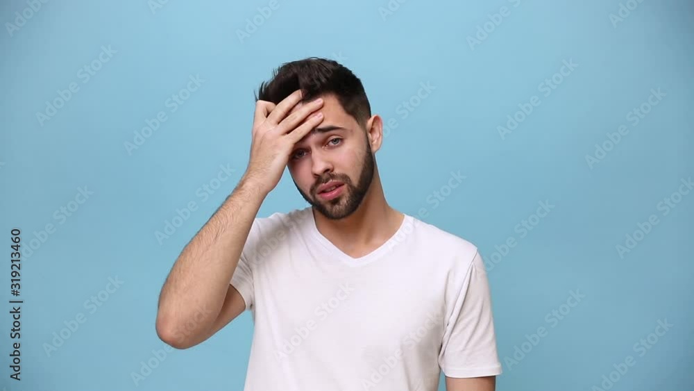 Handsome unshaven bearded young guy 20s wearing white t-shirt isolated over pastel blue background in studio. People sincere emotions lifestyle concept. Looking at camera tired sad put hand on face Handsome unshaven bearded young guy 20s wearing white t-shirt isolated over pastel blue background in studio. People sincere emotions lifestyle concept. Looking at camera tired sad put hand on face