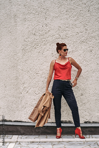 Portrait of a red-haired middle-aged woman in a red blouse and in sunglasses standing against the wall. Cloudy summer day. Close-up.