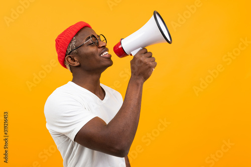 black handsome smiling american man in white t-shirt speaks news through a megaphone on isolated orange background with copy space