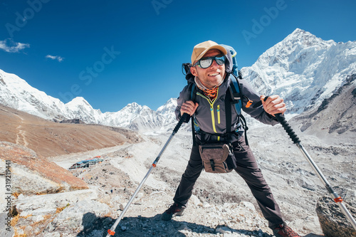 Portrait of smiling Hiker man with Nuptse 7861 m peak and Gorak shep settlement background with trekking poles, UV protecting sunglasses. He enjoying mountain during Everest Base Camp trekking route.