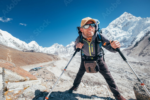 Obraz na plátně Portrait of smiling Hiker man with Nuptse 7861 m peak and Gorak shep settlement background with trekking poles, UV protecting sunglasses