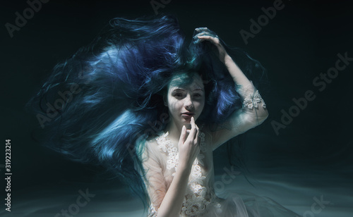 A beautiful girl with blue and long hair swims underwater in the pool in a fluffy white dress. Looks like a nymph or a mermaid
