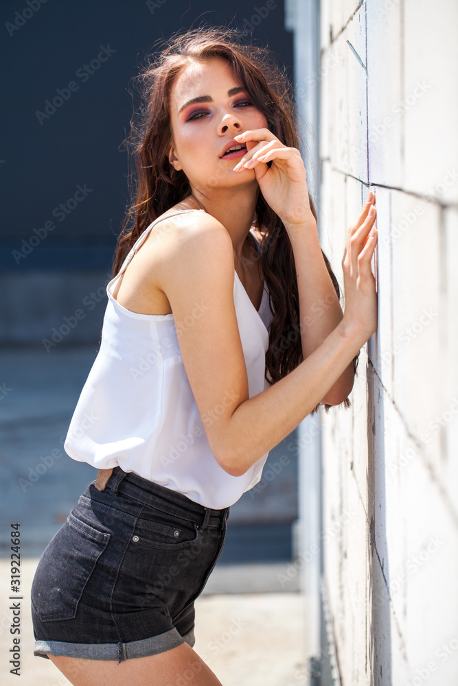 Fototapeta premium Pretty young brunette model in white summer blouse and jeans, summer street outdoors