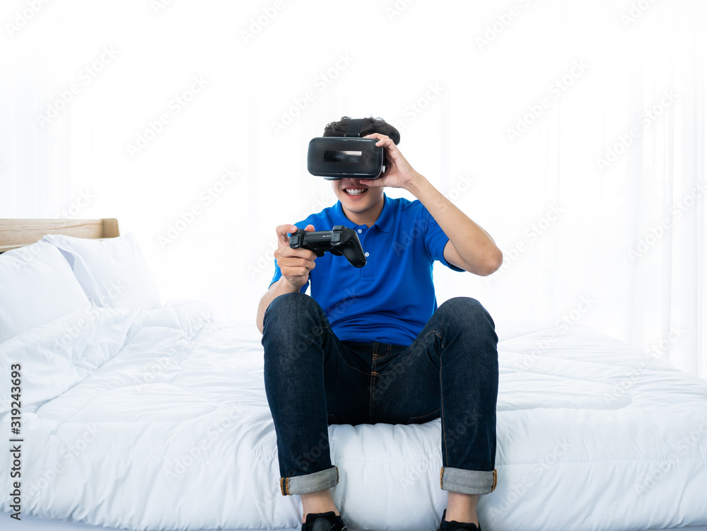 Asian young man enjoy and playing virtual reality goggles simulation digital technology device to connect game online with joystick controller gadget equipment hobbies and leisure at white bedroom Asian young man enjoy and playing virtual reality goggles simulation digital technology device to connect game online with joystick controller gadget equipment hobbies and leisure at white bedroom