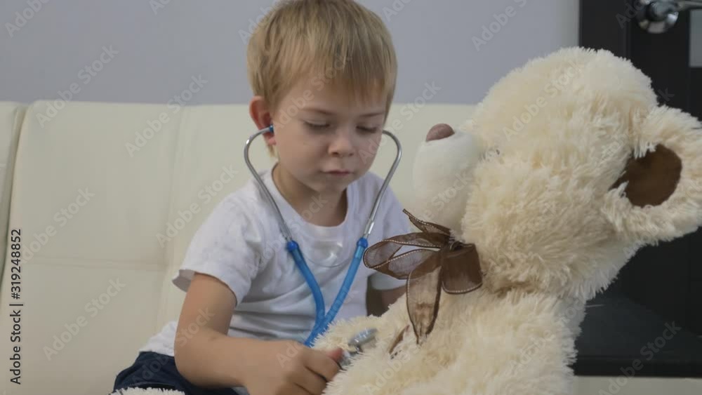 A little boy plays a doctor with a stethoscope and a teddy bear. The boy is sitting on the couch. The concept of children's fantasies and games. Training as a doctor A little boy plays a doctor with a stethoscope and a teddy bear. The boy is sitting on the couch. The concept of children's fantasies and games. Training as a doctor
