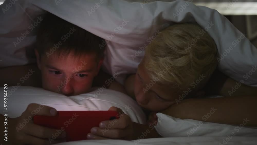 Children lie under a blanket at night and use the game app on their tablet. Children lie under a blanket at night and use the game app on their tablet.