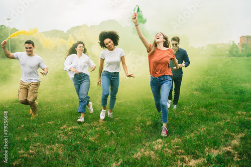Group of five friends runs in a park with two smoke bombs at the park - Millennials have fun together in the summer at sunset