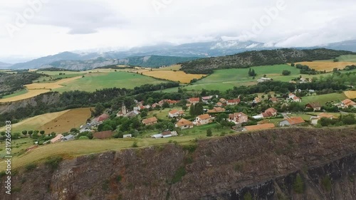 A tiny village in the mountains of French Alps located on cliff. Fields and meadows around, mountain peaks on the horizon. Cozy rural scenery in summer at cloudy day