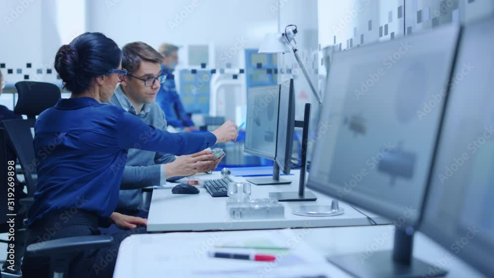 Modern Factory: Male Industrial Engineer Explains to Female Project Supervisor Functions of the Machine Part Comparing it to one on Computer Screen. They use CAD Software for Design, Development Modern Factory: Male Industrial Engineer Explains to Female Project Supervisor Functions of the Machine Part Comparing it to one on Computer Screen. They use CAD Software for Design, Development
