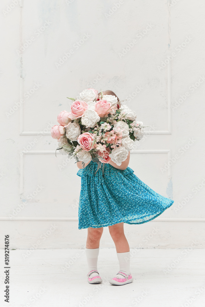 Cute little girl child in a summer dress holds a large bouquet of flowers near a white wall. Studio glamour shot