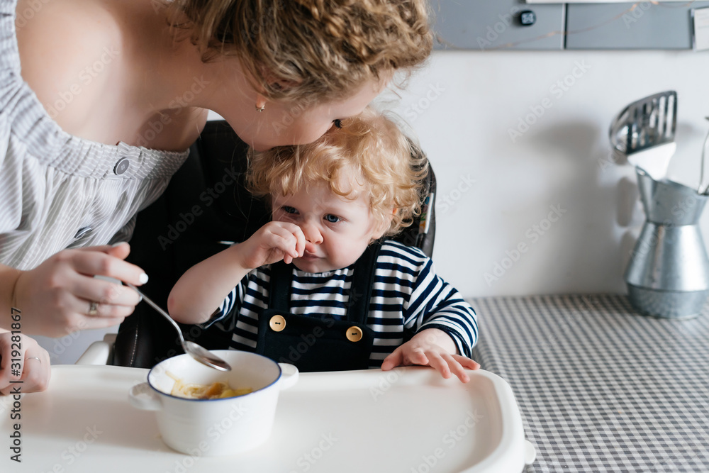 blonde mother feeds the blonde baby with a spoon and gently kisses the top of his head blonde mother feeds the blonde baby with a spoon and gently kisses the top of his head