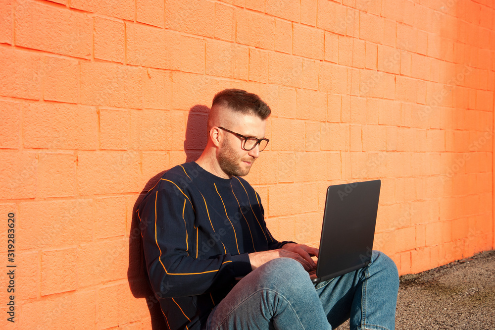 Young caucasian man in casual-wear sitting cross-legged on ground and working on laptop. Freelancer working outside typing on laptop computer. Red wall on the background. Young caucasian man in casual-wear sitting cross-legged on ground and working on laptop. Freelancer working outside typing on laptop computer. Red wall on the background.